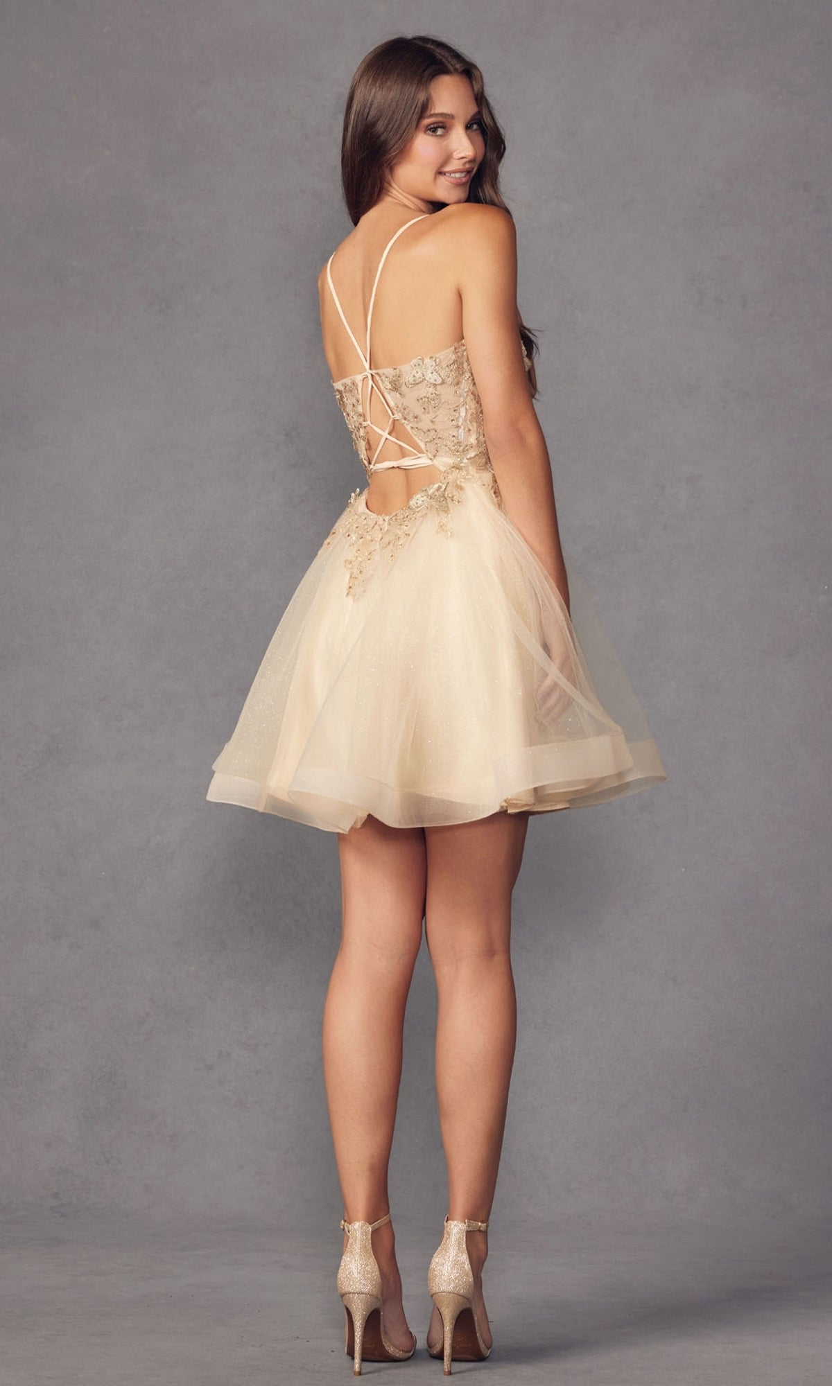 Woman wearing the Short A-Line Butterfly Homecoming Dress 902, a beige tulle babydoll party dress with lace details and crisscross open-back straps, standing and looking over her shoulder.